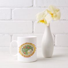 a white vase with flowers in it next to a coffee mug