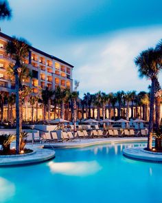 an outdoor swimming pool with chaise lounges and palm trees at dusk in front of a hotel