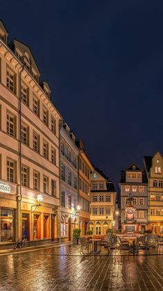 an empty city square at night with tables and chairs on the ground in front of buildings