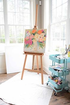 an easel with flowers painted on it in front of a window and a white rug