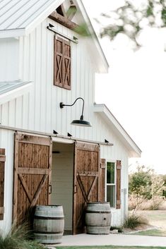 a white barn with wooden doors and two wine barrels in front of it, sitting on the grass