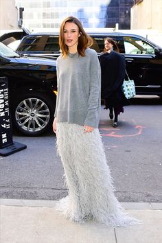 a woman standing in front of a black car wearing a gray sweater and feather skirt