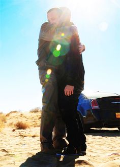 two people are standing in the sand near a car and one person is holding onto another man's back