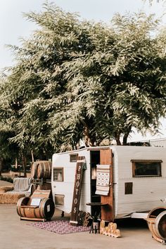 an old camper trailer is parked under a tree with barrels on the ground next to it