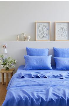 a bed with blue sheets and pillows in a white room next to two pictures on the wall