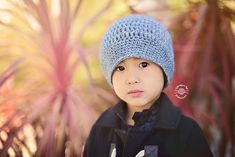 a young boy wearing a blue knitted hat standing in front of some plants and trees