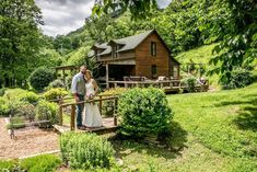 a bride and groom standing on a deck in front of a wooden cabin surrounded by greenery