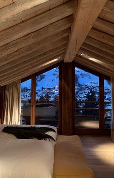 a bed sitting under a wooden ceiling next to two large windows with snow covered mountains in the background