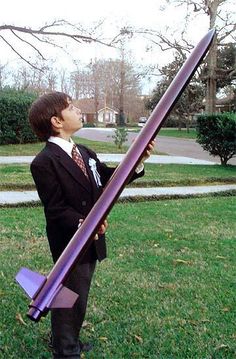 a young boy dressed in a suit and tie holding a giant purple plastic object while standing on the grass