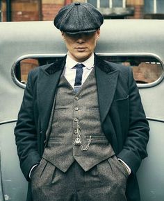 a man in a suit and hat standing next to an old car with his hands in his pockets