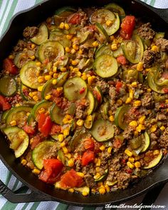 a skillet filled with meat and vegetables on top of a striped table cloth next to a wooden spoon