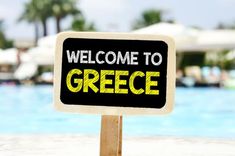 a sign that says welcome to greece in front of a swimming pool with palm trees