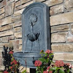 a bird fountain in front of a brick wall with flowers around it and plants growing next to it