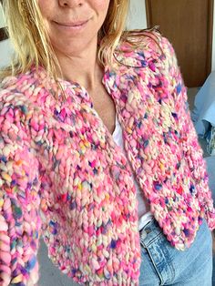 a woman wearing a pink and multicolored knitted cardigan standing in front of a bed