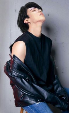 a man sitting on top of a wooden chair wearing a black shirt and leather jacket