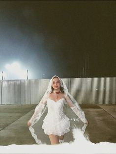 a woman wearing a wedding dress and veil in front of a white fence at night