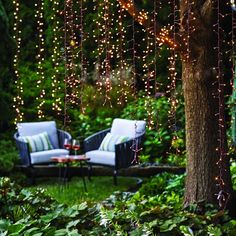 two chairs and a table in the middle of a garden with lights strung from trees