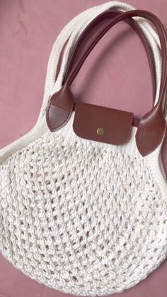 a crocheted white purse with brown leather handles sitting on a pink table top