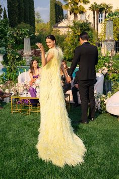 a woman in a long yellow dress standing on top of a lush green field next to a man