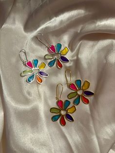 three colorful flower shaped earrings laying on top of a white satin material covered tablecloth