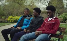 three young men sitting on a bench in a park, one is looking at the camera