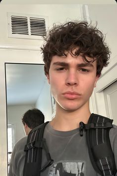 a young man with curly hair wearing a gray shirt and black backpack looking at the camera