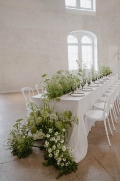 a long table is set with white chairs and flowers in the center, along with greenery