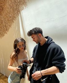 a man standing next to a woman holding a camera in front of a white wall