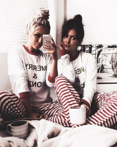 two women in matching pajamas sitting on a bed looking at their cell phones and drinking coffee