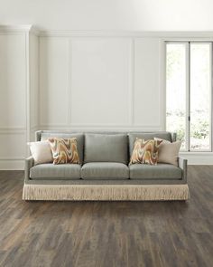 a living room with hardwood floors and white walls, including a gray couch that has fringe trim around the edges