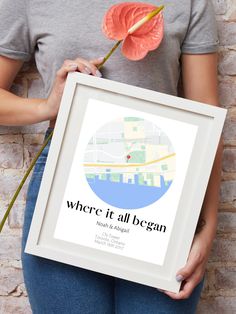 a woman holding up a framed poster with a flower in front of her face and the words where it all begun