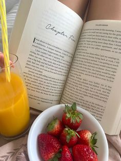 a bowl of strawberries next to an open book and a glass of orange juice