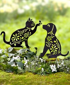 two black cats standing next to each other in front of flowers and plants on the ground