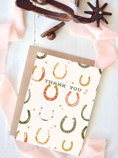 a thank you card with horseshoes and pink ribbon on the table next to it