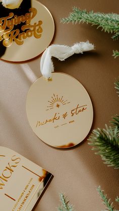 two gold and white wedding tags sitting on top of a table next to pine branches