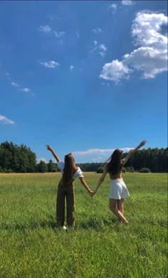 two young women are holding hands in the middle of a grassy field with trees and blue sky behind them