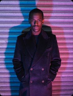 a man standing in front of a wall wearing a purple coat and black pants with his hands in his pockets