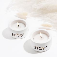 two white candles with hebrew writing on them sitting in front of a fluffy white background