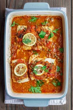 a casserole dish filled with fish, tomatoes and lemons on top of a wooden table