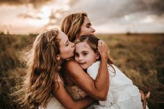 three girls hugging each other in a field with the sun shining through clouds behind them