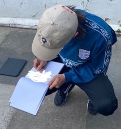 a man sitting on the ground drawing something with white paper and a baseball cap over his head