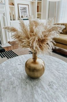 a white marble table with a gold vase filled with dry grass on it and a couch in the background