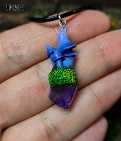 a hand holding a purple and green necklace with a blue flower on it's end