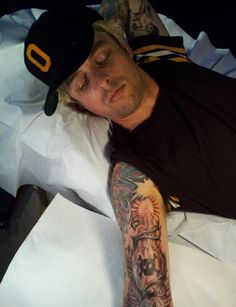 a man laying in bed with tattoos on his arm and chest, wearing a baseball cap