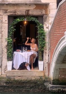 a woman sitting at a table in front of a window eating food and drinking wine