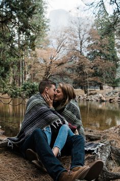 a man and woman sitting next to each other in front of a lake with trees