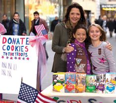 two girls and an adult stand behind a table with books on it in the street