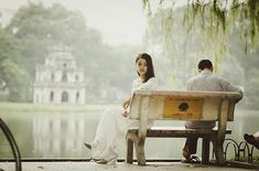 a man and woman sitting on a bench next to a body of water with trees in the background