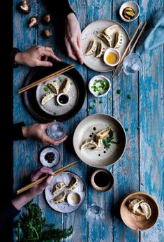 two people eating dumplings with chopsticks on a blue wooden table, surrounded by other dishes