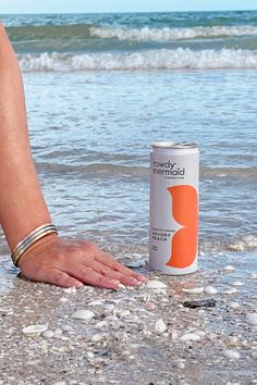 a woman's hand reaching for a can of soda on the beach next to the ocean